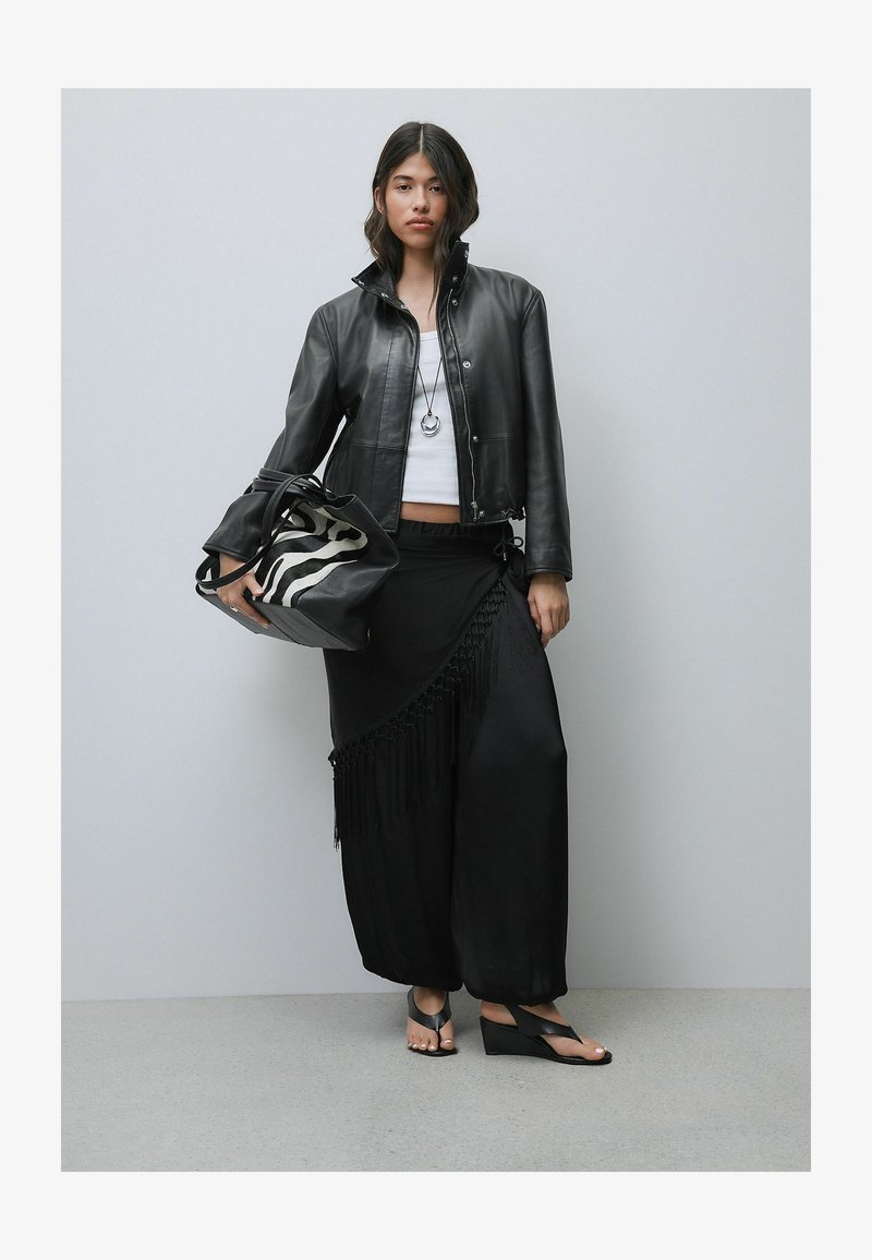Femme portant une veste en cuir noire, un haut blanc, un pantalon large noir avec des franges, tenant un sac à motif zèbre, et portant des sandales compensées noires.