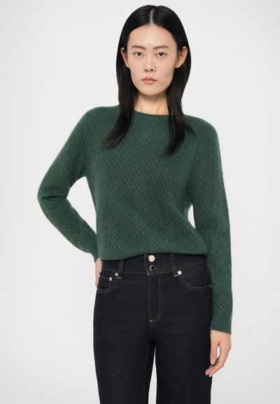 Green knitted sweater with a textured pattern, round neckline, and full sleeves, paired with dark denim jeans featuring front pockets and stitching.