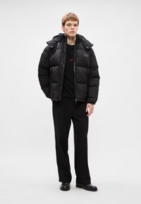 Crna puffer jakna s patentnim zatvaračem i odvojivim kapuljačom, nosi se preko crne dukserice. Model nosi crne hlače i cipele.