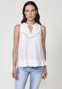 Top bianco senza maniche con scollatura a onda, dettagli in pizzo e orlo a volantino svasato. Tessuto leggero e testurizzato con una vestibilità ampia.