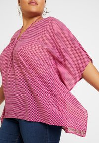 Haut ample à manches courtes rose avec motifs géométriques blancs. Dispose d'un col en V et d'un tissu léger et texturé.