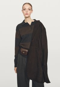 Brązowy sweter w paski z kołnierzykiem, brązowy zamszowy płaszcz zarzucony na ramiona, szare spodnie w kant oraz mała brązowa torba na pasku.