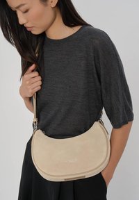 Borsa a tracolla in camoscio beige con design curvo, dettagli in argento e tracolla lunga, indossata con una maglia grigia dalla vestibilità morbida.