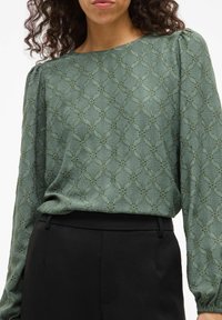 Zelená krajková blůza s dlouhými rukávy, kulatým výstřihem a texturovaným vzorem. V kombinaci s černými kalhotami.