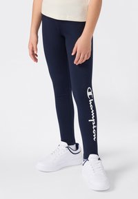 Granatowe legginsy wykonane z elastycznego materiału, z dużym białym logo "Champion" na lewym udzie. Noszone z białymi sneakersami.