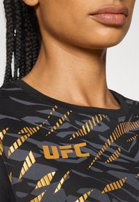 Svart treningstrøye med teksturert mønster i grått og gull, med "UFC"-logo i gull på forsiden, rund hals.