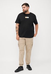 Czarny bawełniany t-shirt z prostokątnym logo, beżowe spodnie cargo z bocznymi kieszeniami i czarne trampki na sznurowadła na jednolitym tle.