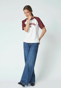 T-shirt bianca con maniche raglan bordeaux e stampa "COUTURE". Abbinata a jeans in denim blu a gamba larga. Design semplice e casual.