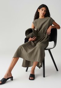 Olivgrünes, knöchellanges Kleid mit kurzen Ärmeln und elastischem Bund, kombiniert mit schwarzen Riemchenschuhen, sitzt auf einem schwarzen Stuhl.