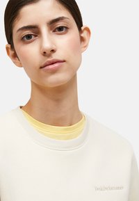 Felpa beige chiaro con colletto a coste, caratterizzata da un delicato ricamo del logo. Indossata sopra una t-shirt gialla, che mette in risalto le texture sovrapposte.