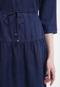 Abito blu navy realizzato in tessuto morbido, con vita aderente e fiocco, maniche a sbuffo e gonna arricciata con bottoni decorativi.