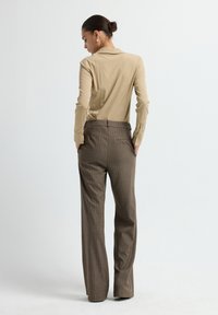 Vrouw staand met handen in de zakken, gekleed in een beige langharige shirt en bruine geruite wijde broeken tegen een witte achtergrond.