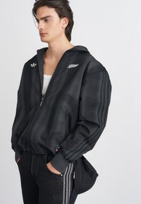 Casaco bomber preto com padrão xadrez, detalhes em prata e fecho de correr na frente. Apresenta dois bolsos laterais e logos bordados no peito.