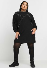 Abito nero a maglia con maniche lunghe, caratterizzato da un motivo a incrocio di borchie argentate. L'abito ha una vestibilità ampia e arriva a metà coscia.
