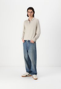 Lys beige langærmet polo med lynlås i halsen, parret med løse blå jeans og hvide sneakers, stående foran en ensfarvet baggrund.
