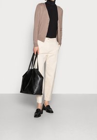 Person som bär en beige kavaj, svart polotröja, krämfärgade byxor och svarta loafers, som håller i en stor svart läderhandväska.