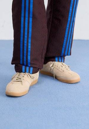 Baskets en daim beige avec semelles en gomme portées avec un pantalon marron foncé à rayures verticales bleues sur un tapis bleu clair.