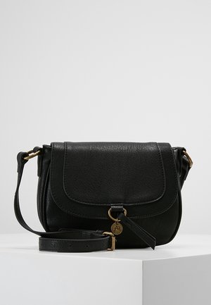 Sac bandoulière en cuir noir avec fermeture à rabat, sangle ajustable et petit pendentif rond doré à l'avant, présenté sur un piédestal blanc.