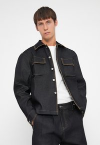 Donkerdenimjack met lichtbruine stiksels, voorzien van een puntige kraag, twee borstzakken en drukknoopsluiting. G gedragen over een wit shirt.