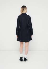 Mørk denim kåpekjole med en figursydd silhuett, lange ermer og synlige sømdetaljer. Kantlinjen er utsvingt, og lommene er til stede.