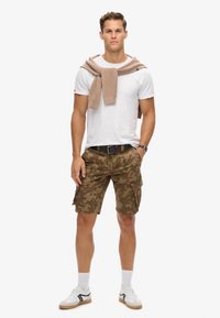 Witte T-shirt, beige sweater over de schouders, camouflage cargo shorts, zwarte riem, witte sokken met bruine en zwarte sneakers, staande pose.