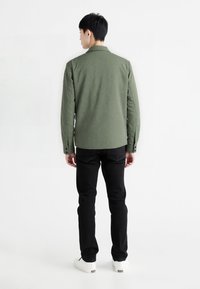 Olivengrøn skjorte med et struktureret mønster, knapmanchetter og krave, parret med sorte jeans og hvide sneakers. Set fra ryggen.