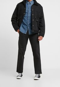 Svart denimjacka med en shearlingkrage, bärs över en blå skjorta med knappar, kombinerad med svarta byxor och svarta sneakers.