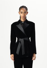 Black velvet blazer with satin lapels, wrap design, and a matching fabric belt. Features two front pockets and a tailored fit.