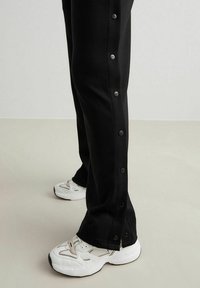 Sorte tapered bukser med trykknapper i siden, lavet af blødt materiale, parret med hvide sporty sko med net- og teksturdetaljer.