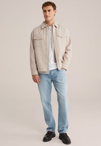Beige knoopjack met twee bovenzakken, gelaagd over een wit shirt en gecombineerd met lichtblauwe jeans en zwarte schoenen.