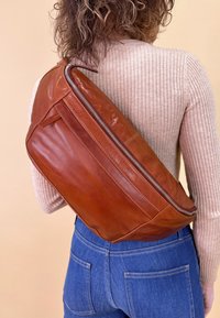 Rudas odinė petinė rankinė su lygiu paviršiumi, turinti įstrižą užtrauktuką ir plokščią struktūrą. Nešiojama per petį, derinama su džinsais.