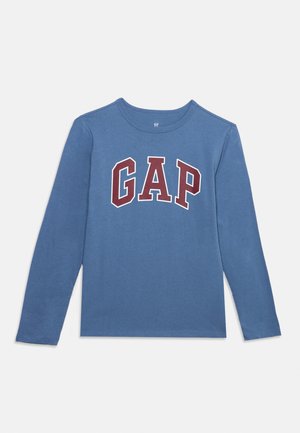Синя риза с дълъг ръкав с кръгло деколте и големи червени и бели "GAP" букви на гърдите върху бял фон.