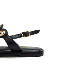 Sandal de cuero negro con suela plana, que presenta un acento circular en tono dorado y tira ajustable en el tobillo. Diseño simple, textura suave.