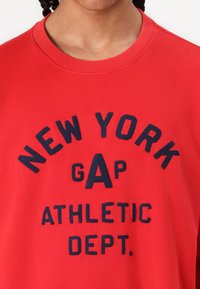 Sudadera de algodón rojo con texto en azul marino que dice "DEPARTAMENTO ATLÉTICO GAP DE NUEVA YORK". Cuello acanalado y corte relajado.