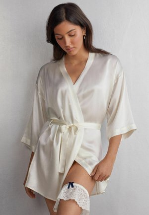 Satin kimono robe i blød elfenben med bælte i taljen og brede ærmer. Med en blondebundet støttestrømpe med en blå sløjfe på låret. Glat tekstur.