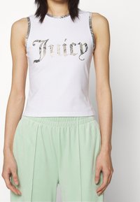 Bílé bezramínkové tričko s nápisem "Juicy" v metalických hadích písmenech a stříbrným lemováním. V páru se světle zelenými kalhotami s elastickým pasem.