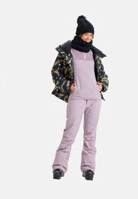 Veste de ski multicolore avec un motif géométrique, haut en polaire violet et pantalon de neige violet clair. Un bonnet et une écharpe en tricot noirs complètent la tenue.