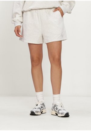 Persoon draagt lichtgrijze shorts, witte sokken en zilver-witte New Balance sneakers, staande tegen een effen achtergrond.