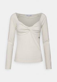 Top beige de manga larga con un diseño frontal retorcido y escote redondeado. Presenta un pequeño logo bordado en el lado inferior. Textura de tela suave.