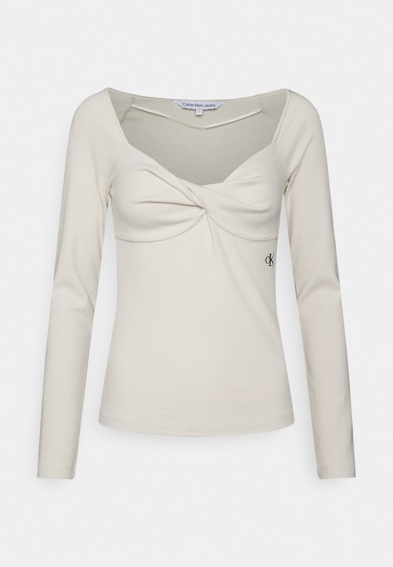 Top beige de manga larga con un diseño frontal retorcido y escote redondeado. Presenta un pequeño logo bordado en el lado inferior. Textura de tela suave.