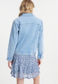 Giacca in denim azzurro chiaro con taglio classico, dotata di bottoni metallici e dettagli di cucitura, abbinata a un vestito a strati con motivo floreale.