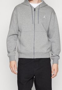 Sudadera gris con cremallera hecha de un tejido suave, presenta un bolsillo tipo canguro, puños acanalados y un pequeño logo en el pecho izquierdo.