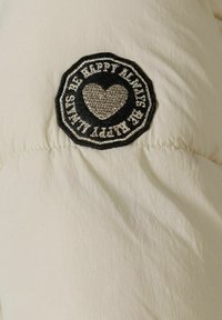 Le tissu de la doudoune beige présente un patch circulaire noir proéminent avec du texte brodé blanc et argent ainsi qu'un motif en forme de cœur.