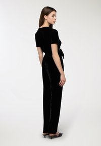 Czarny welurowy kombinezon z krótkimi rękawami, zawiązywaniem w talii i prostymi nogawkami. Gładka tekstura i subtelny połysk.