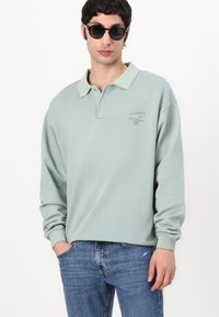 Light green long-sleeve polo shirt with a collar and a subtle graphic logo. Paired with blue jeans, model wears dark sunglasses.