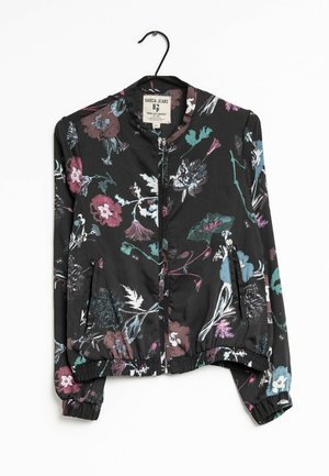 Czarna lekka kurtka z wielokolorowym kwiatowym nadrukiem, z przodu na zamek błyskawiczny, z elastycznymi mankietami i ściągaczem u dołu, wisząca na czarnym wieszaku.