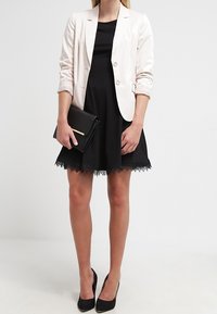 Blazer rosa chiaro sopra un abito nero con bordi in pizzo, abbinato a tacchi neri e una pochette. Tessuto liscio, vestibilità su misura e design elegante.