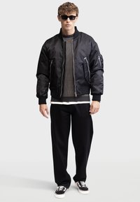 Zwarte bomberjack met ritszakken, geribbelde manchetten en een gemêleerd grijs trui eronder. Losse zwarte broek en zwarte sneakers maken de look compleet.