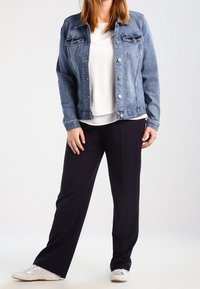 Veste en jean bleu clair avec fermeture à boutons, deux poches poitrine, portée sur un haut blanc. Pantalon noir à jambes larges et baskets blanches.
