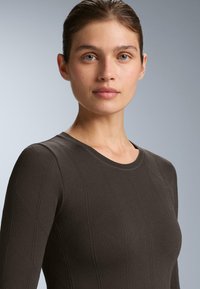 Žena s viazanými vlasmi, oblečená v tmavohnedom dlhom rukáve s textúrou, stojí čelom voči jednoduchému šedému pozadiu.